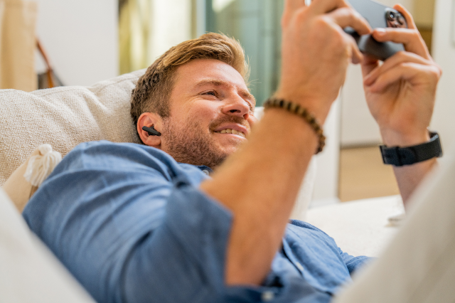 Man on couch wearing Poly earbud headset while looking at his phone