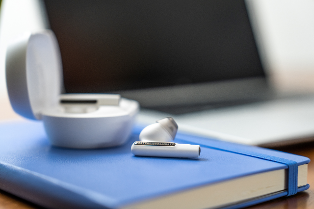 Close up of white Voyager earbuds from Poly on a blue notebook, charging case out of focus in background